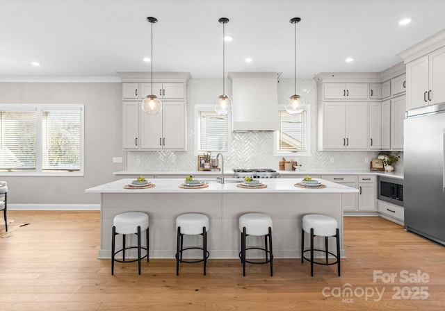 kitchen featuring a breakfast bar area, custom exhaust hood, light countertops, an island with sink, and built in appliances