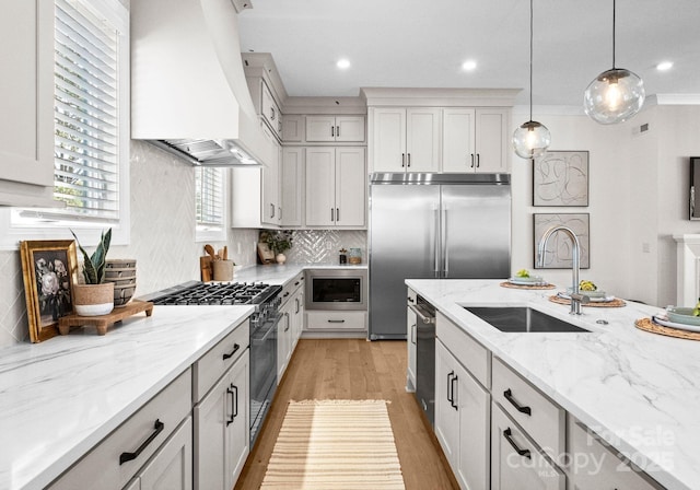 kitchen featuring custom range hood, light stone counters, decorative light fixtures, built in appliances, and a sink