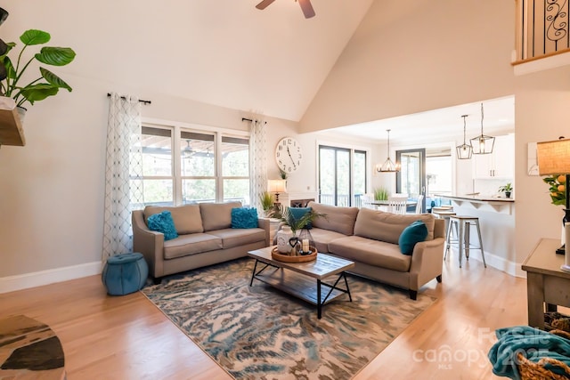 living room featuring ceiling fan with notable chandelier, high vaulted ceiling, light wood-type flooring, and baseboards