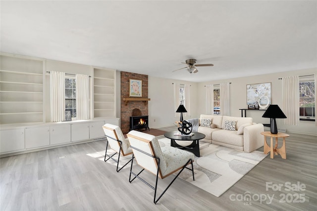 living area with built in shelves, light wood-style flooring, a ceiling fan, baseboards, and a brick fireplace