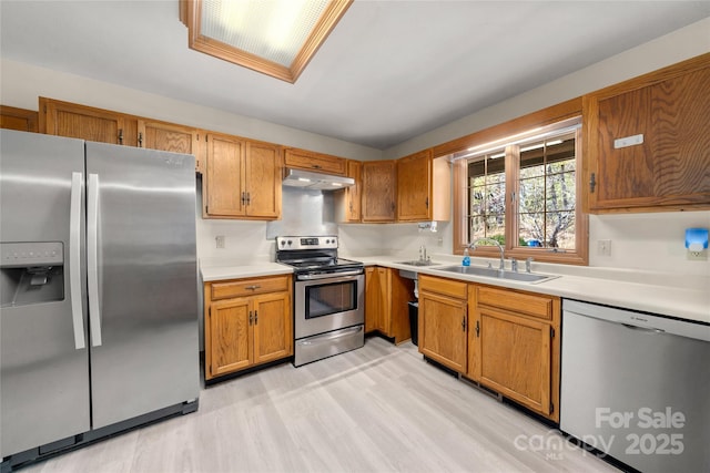 kitchen with brown cabinets, stainless steel appliances, light countertops, under cabinet range hood, and a sink