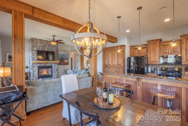 dining room with a stone fireplace, recessed lighting, ceiling fan with notable chandelier, dark wood-type flooring, and beamed ceiling