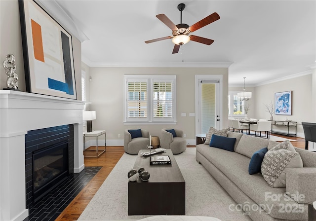 living room featuring wood finished floors, baseboards, a fireplace with flush hearth, ornamental molding, and ceiling fan with notable chandelier