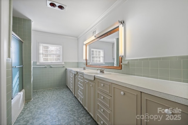 full bath featuring double vanity, tile walls, wainscoting, crown molding, and a sink