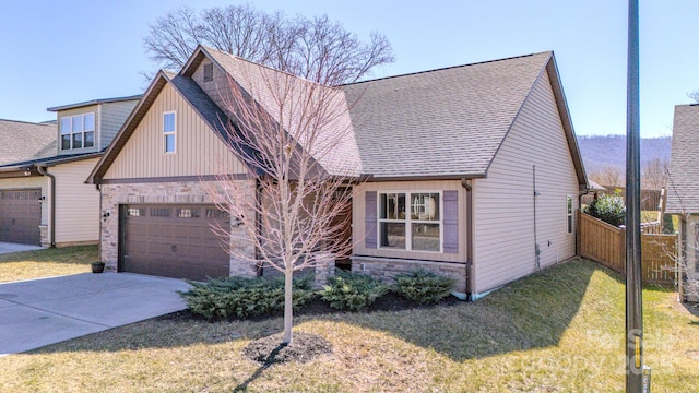 view of front of house featuring a shingled roof, concrete driveway, stone siding, fence, and a front yard