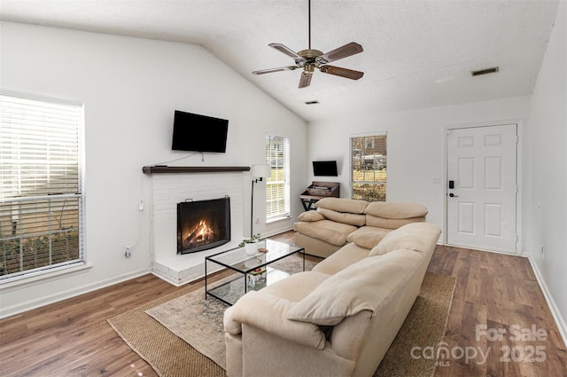 living area featuring lofted ceiling, wood finished floors, visible vents, a ceiling fan, and a brick fireplace