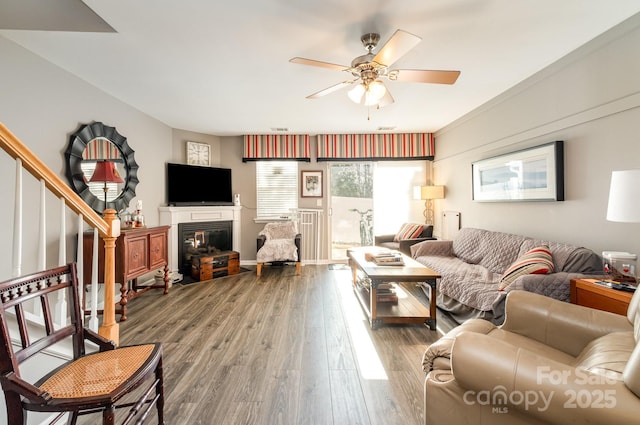 living area with a ceiling fan, a glass covered fireplace, stairway, and wood finished floors