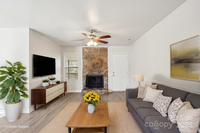 living area featuring light wood-style floors, ceiling fan, baseboards, and a stone fireplace