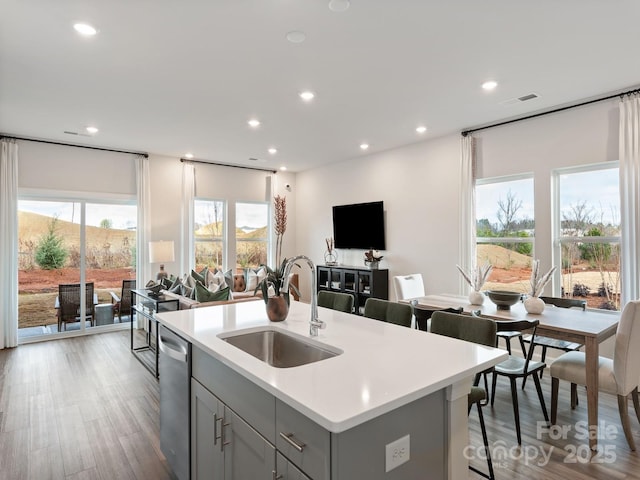 kitchen featuring a center island with sink, dishwasher, light countertops, gray cabinetry, and a sink