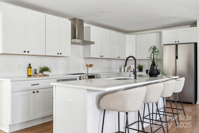 kitchen featuring a kitchen island with sink, stainless steel appliances, a sink, white cabinets, and wall chimney exhaust hood