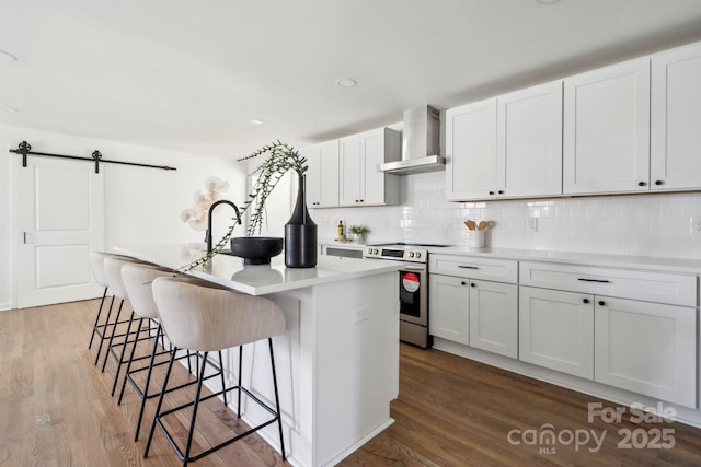 kitchen featuring a barn door, a center island with sink, electric stove, wall chimney range hood, and backsplash