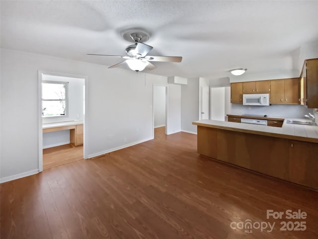 kitchen featuring baseboards, brown cabinetry, white microwave, dark wood-style floors, and a peninsula