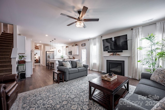 living room featuring recessed lighting, a ceiling fan, stairs, dark wood-style floors, and a glass covered fireplace