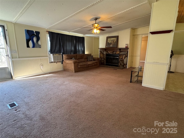 living room with visible vents, a ceiling fan, a wainscoted wall, carpet flooring, and a stone fireplace