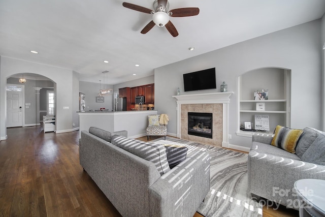 living room with baseboards, built in features, arched walkways, a tile fireplace, and dark wood-style floors