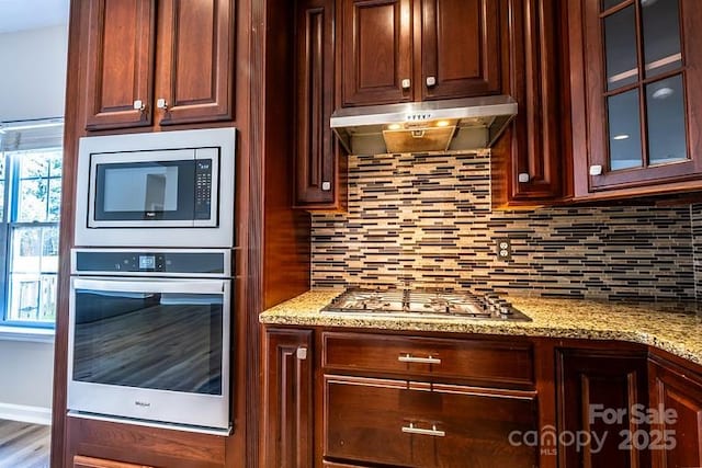 kitchen with light stone counters, stainless steel appliances, decorative backsplash, glass insert cabinets, and under cabinet range hood