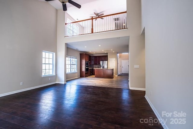 unfurnished living room with ceiling fan, dark wood-style flooring, a towering ceiling, and baseboards