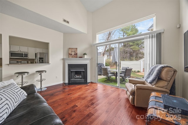 living room featuring a fireplace with raised hearth, a towering ceiling, visible vents, baseboards, and wood-type flooring