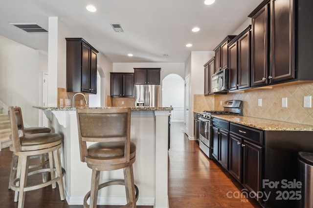 kitchen featuring arched walkways, visible vents, dark brown cabinets, appliances with stainless steel finishes, and light stone countertops