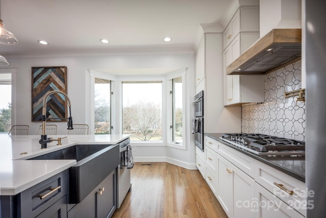 kitchen with light wood-style flooring, stainless steel appliances, crown molding, wall chimney range hood, and a sink