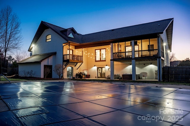 back of house at dusk featuring a balcony, a patio area, and fence