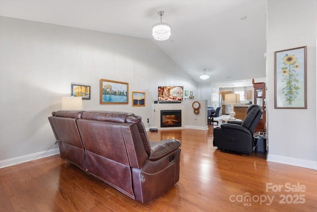 living room featuring a glass covered fireplace, high vaulted ceiling, baseboards, and wood finished floors