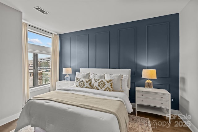 bedroom with visible vents, a decorative wall, baseboards, and wood finished floors