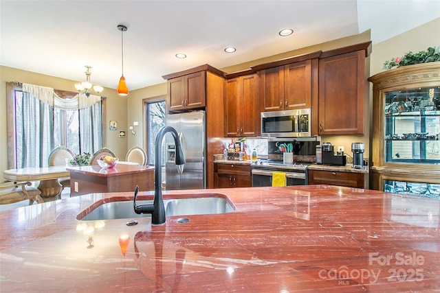 kitchen featuring brown cabinetry, dark stone countertops, decorative light fixtures, stainless steel appliances, and a sink