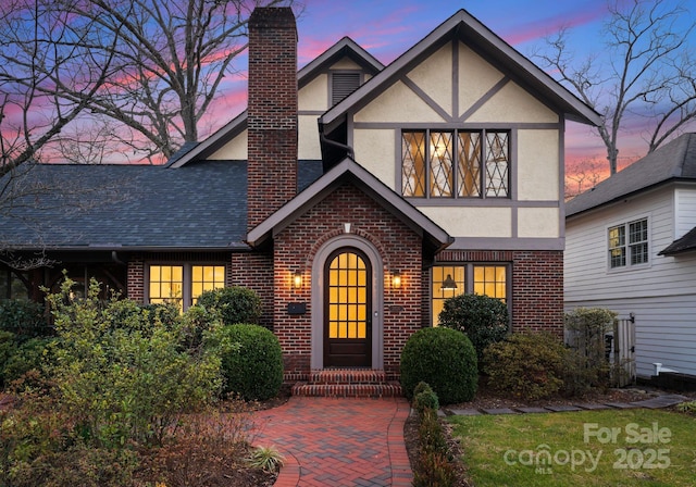 tudor-style house featuring brick siding, roof with shingles, a chimney, and stucco siding