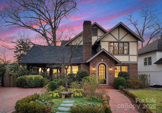 tudor-style house featuring a shingled roof, brick siding, a chimney, and stucco siding