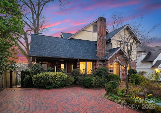 english style home featuring brick siding, a shingled roof, a chimney, fence, and stucco siding