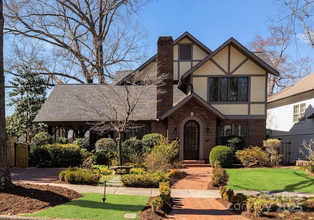 tudor home with a shingled roof, stucco siding, a chimney, a front lawn, and brick siding