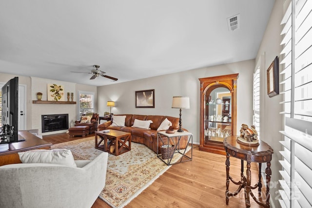 living room with light wood finished floors, a glass covered fireplace, visible vents, and a ceiling fan