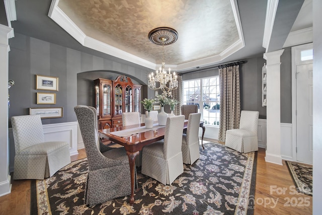 dining space with a wainscoted wall, wood finished floors, a raised ceiling, and decorative columns