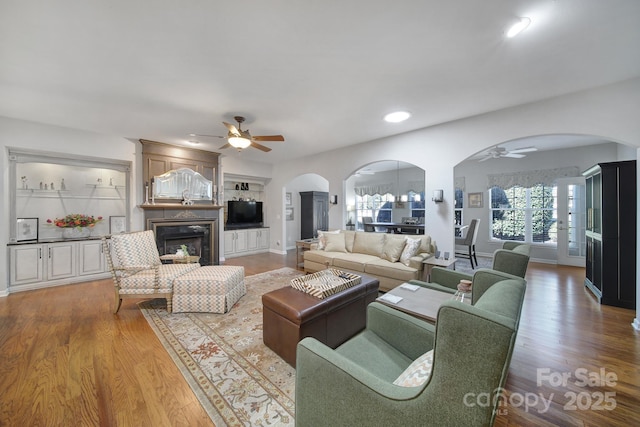 living room featuring arched walkways, a ceiling fan, a glass covered fireplace, wood finished floors, and baseboards