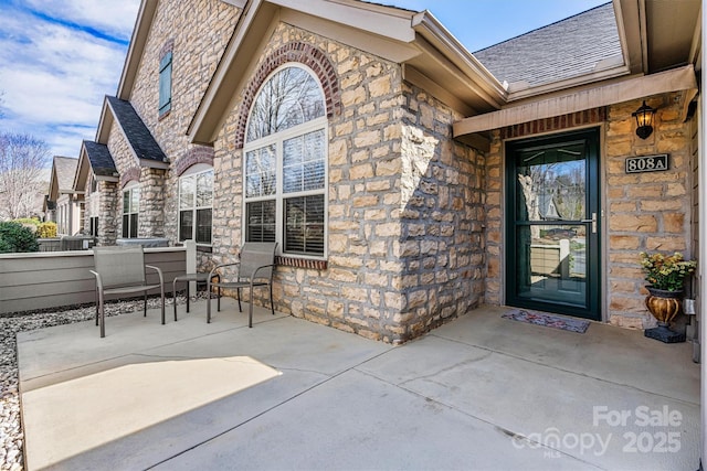 doorway to property featuring stone siding, a shingled roof, and a patio