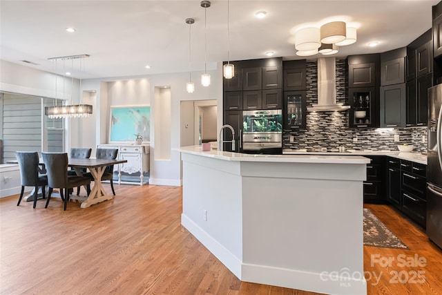 kitchen with light countertops, appliances with stainless steel finishes, wall chimney exhaust hood, light wood finished floors, and tasteful backsplash