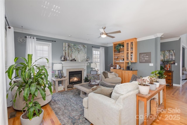 living area featuring light wood-style floors, ornamental molding, a ceiling fan, and a glass covered fireplace