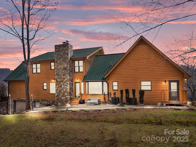 back of property at dusk featuring central air condition unit, a garage, entry steps, metal roof, and a chimney