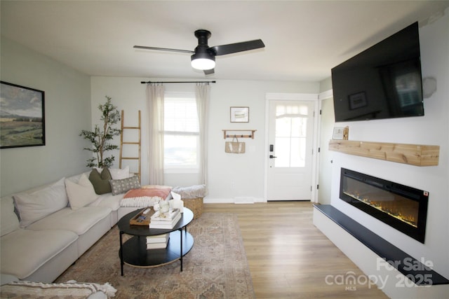 living room featuring baseboards, a glass covered fireplace, light wood-style flooring, and a ceiling fan