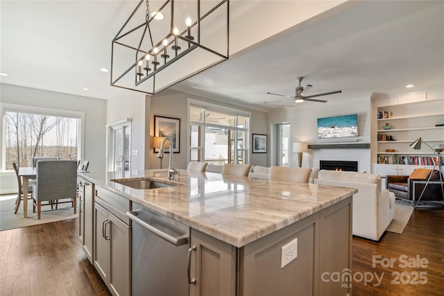 kitchen featuring a large fireplace, an island with sink, dark wood-style floors, stainless steel dishwasher, and a sink