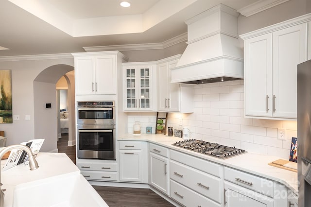 kitchen featuring arched walkways, stainless steel appliances, a sink, a tray ceiling, and custom range hood