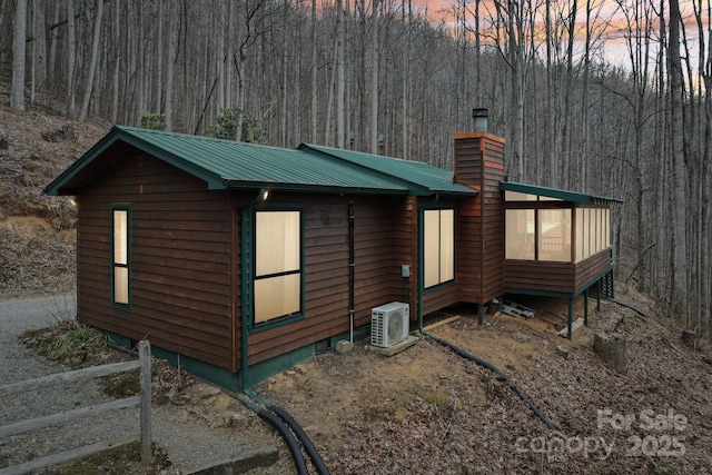 property exterior at dusk with ac unit, metal roof, a chimney, and a wooded view