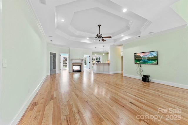 unfurnished living room with ceiling fan, a multi sided fireplace, ornamental molding, light wood-type flooring, and a tray ceiling