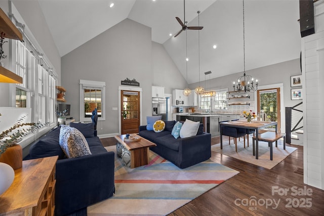 living room featuring baseboards, high vaulted ceiling, dark wood-type flooring, and ceiling fan with notable chandelier