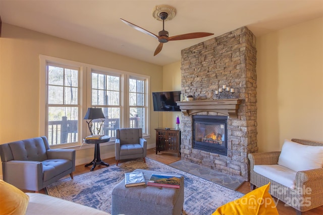 living area with ceiling fan, a stone fireplace, baseboards, and wood finished floors