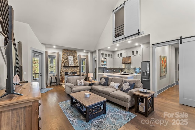 living area with a barn door, high vaulted ceiling, wood finished floors, and a stone fireplace