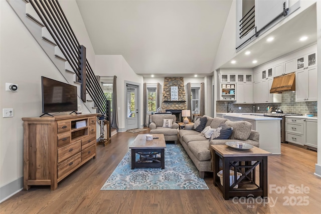 living area featuring high vaulted ceiling, a stone fireplace, wood finished floors, baseboards, and stairway