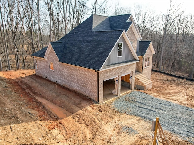 view of property exterior with gravel driveway, brick siding, roof with shingles, an attached garage, and board and batten siding