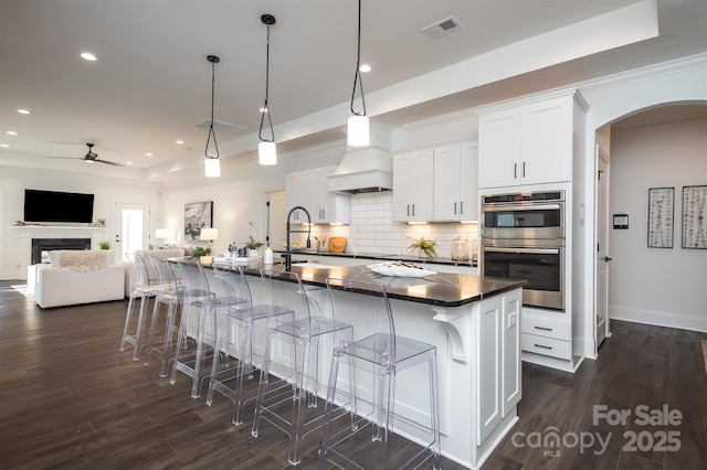 kitchen with arched walkways, double oven, premium range hood, visible vents, and dark countertops